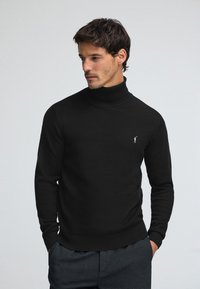 Svart polotröja i stickat material med åtsittande långärmar och en diskret logga. Mjuk textur, minimalistisk design.