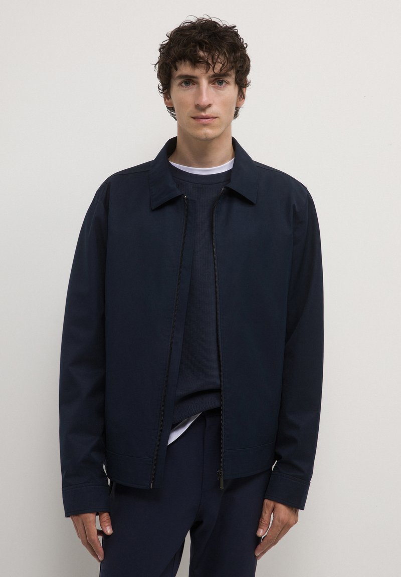 Giacca navy con zip frontale, colletto piatto e maniche lunghe. Indossata sopra un maglione navy a tessuto strutturato. Tessuto liscio, dettagli minimi.