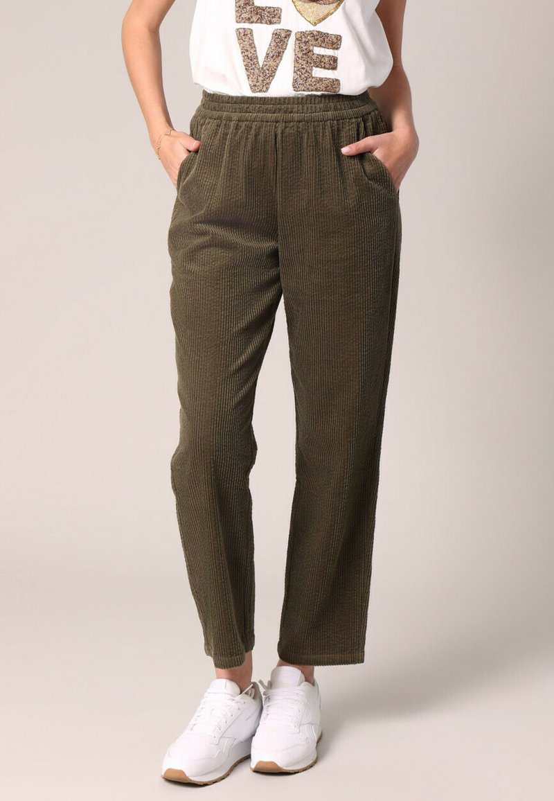 Olijfgroene, ribbelige corduroy broek met een elastische tailleband, voorzien van zijzakken en een taps toelopend ontwerp. Gecombineerd met witte sneakers.