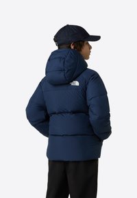 Tumesinine puffer-jope kapuutsiga, horisontaalse õmbluse ja seljal oleva valge logoga. Sobitatud tumeda teksakmütsiga.