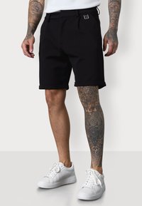 Black shorts with a pleated design and rolled cuffs, featuring a small logo detail. Worn with white sneakers and a visible tattoo.