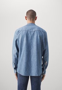 Blå denimskjorta med lång ärm, rundad nederkant, knäppta manschetter och en liten patchdetalj på sidan; mjuk textur och avslappnad passform.