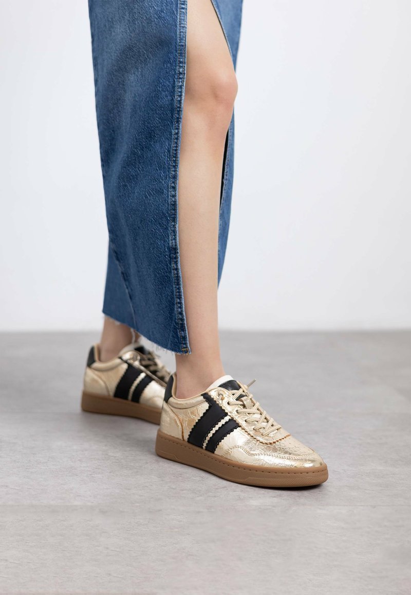 Zapatillas metálicas doradas con rayas negras, Upper texturizado y suelas de goma marrón, combinadas con una falda de mezclilla con una abertura lateral.