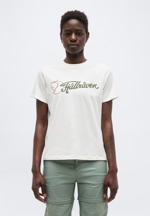 Person med kort hår iført en hvid T-shirt med Fjällräven-logo og lysegrønne cargobukser, stående mod en ensfarvet hvid baggrund.