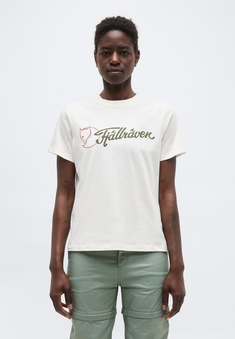 Άτομο με κοντά μαλλιά που φοράει άσπρο t-shirt με το λογότυπο Fjällräven και ανοιχτοπράσινο παντελόνι cargo, στέκεται μπροστά σε λευκό απλό φόντο.