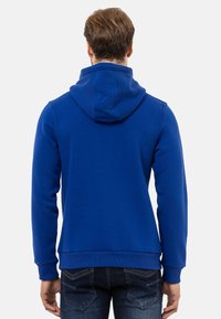 Blaues Hoodie aus weichem, strukturiertem Stoff mit einer Kordelzugkapuze, gerippten Bündchen und geradem Saum, kombiniert mit dunklen Jeans.