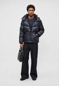 Casaco acolchoado azul-marinho com capuz, design matelassê e fecho de correr frontal, usado sobre um suéter azul-marinho e jeans escuros. Segura uma mala preta.