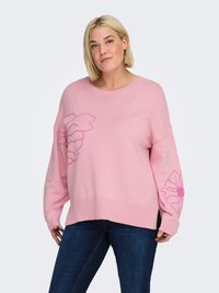 Pink knitted sweater with long sleeves, featuring a floral design on the front and left sleeve. Ribbed hem. Paired with dark blue jeans.