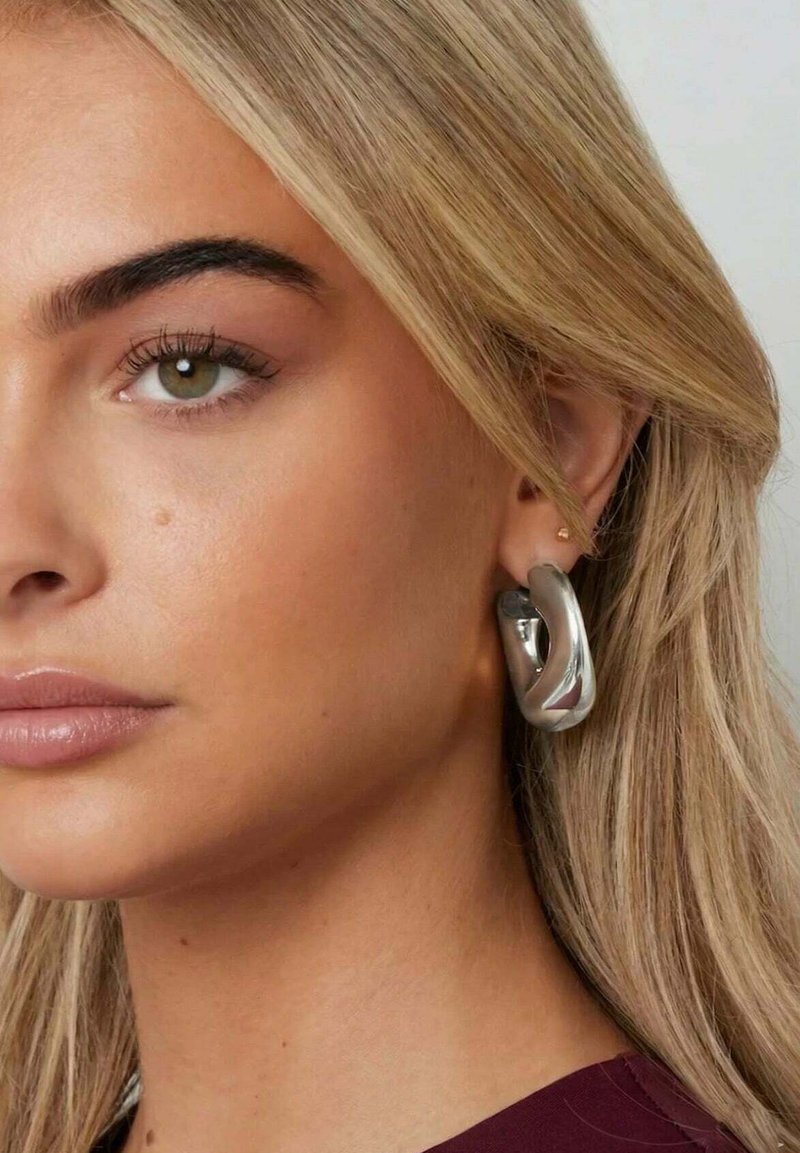 Woman with light blonde hair wearing large, shiny silver hoop earrings and a burgundy top, looking slightly to the left.