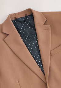 Blazer marron avec doublure intérieure noire à pois blancs et une boucle de chaîne noire près du col sur un fond clair.