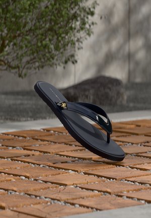 Sort klipklap-sandal med en guldemblem på remmen, placeret på et mønstret trægulv med grønt løv og en grå væg i baggrunden.