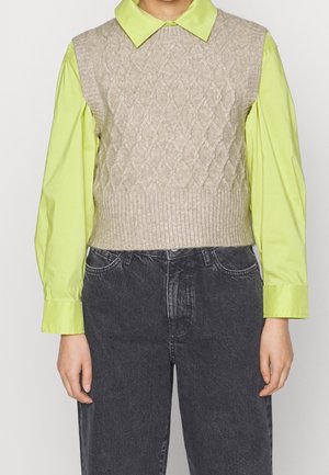 Persona que lleva una camisa de manga larga color verde lima con cuello, debajo de un chaleco sin mangas de punto grueso color beige y vaqueros negros desgastados.