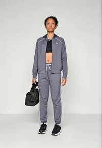 Grå treningsjakke med glidelås og hvite detaljer, matchende grå joggebukser med sidestipler, svart sportsbh og svart treningsbag.