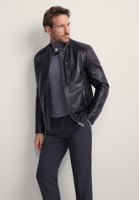 Czarna skórzana kurtka z wysokim kołnierzem, zapinana na zamek z przodu oraz z zamkami błyskawicznymi w kieszeniach, zestawiona z dopasowanym szarym swetrem i ciemnoszarymi spodniami.