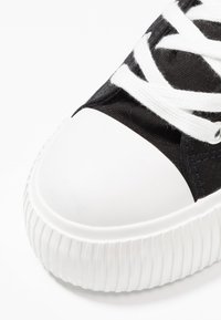 Sort lærreds-sneaker med hvid gummistskærm foran og tekstureret hvid sål. Snørebåndene er hvide og flade.