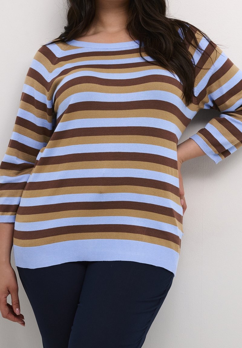Femme portant un pull rayé bleu clair, marron et beige à manches trois-quarts et un pantalon foncé, debout avec une main sur la hanche.