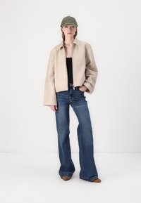 Beige skinnjakke med brede ermer, kombinert med mørkeblå bredbente jeans og en grønn caps. Modellen står foran en hvit bakgrunn.