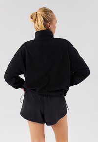Černý fleecový pulovr s vysokým límcem a volným střihem. Má dlouhé rukávy a nastavitelný lem se šňůrkou. Nošený s černými šortkami.