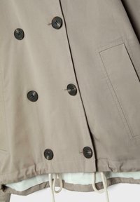 Beige dobbeltradet frakke med mørke knapper, synlig lomme og hvid foring med snøre i kanten.