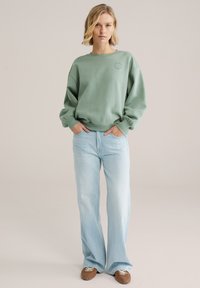 Mintgroene sweatshirt met doorgezakte schouders, geribbelde manchetten en een ronde logo. Gecombineerd met lichtblauwe broek met brede pijpen en bruine sneakers.