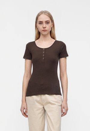 Jeune femme aux longs cheveux blonds porte un haut moulant à manches courtes côtelé marron foncé et un pantalon beige clair, debout devant un fond uni.