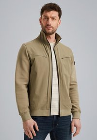 Giacca zip-up verde oliva con colletto alto, due tasche sul petto e polsini a costine. Abbinata a una camicia a righe chiare e jeans scuri.