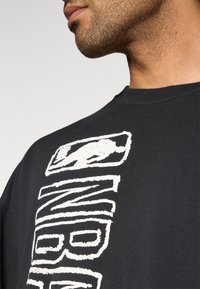 Μαύρο βαμβακερό μπλουζάκι με έντονο λευκό γραφικό κείμενο στο πλάι, με στρογγυλή λαιμόκοψη και κοντά μανίκια. Ορατή υφή υφάσματος.