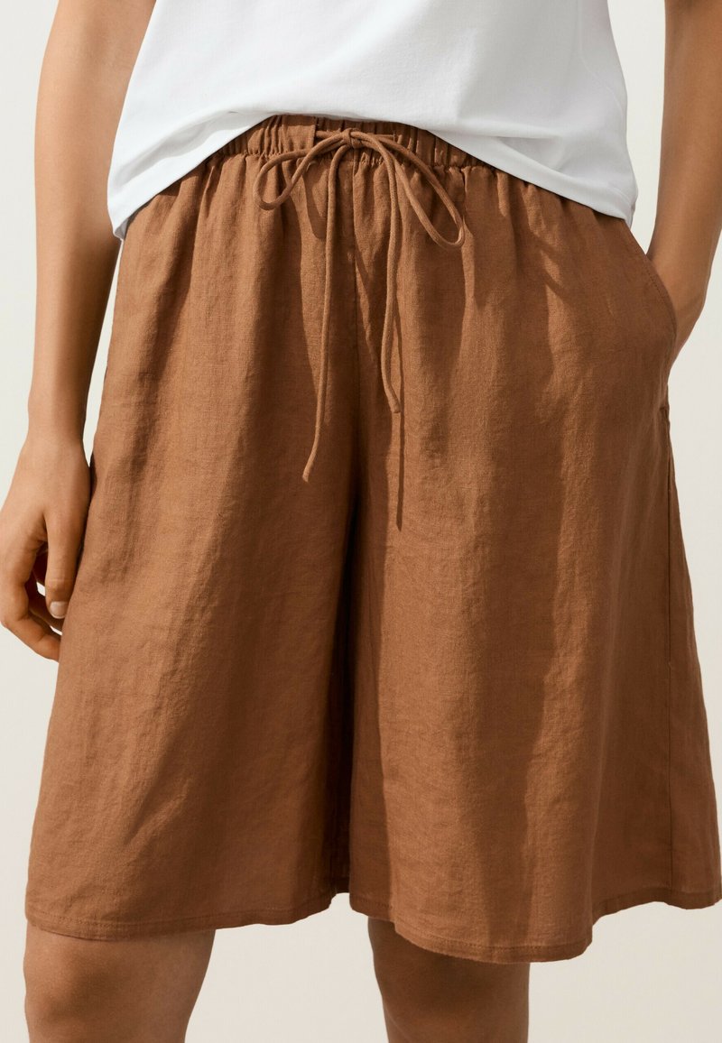Shorts marron en lin longueur genou avec une ceinture élastique et un cordon, portés avec un haut blanc, mains détendues sur les côtés.