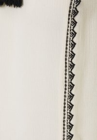 Tissu couleur crème présentant une broderie noire élaborée le long du bord, avec un motif zigzag décoratif et une surface texturée.