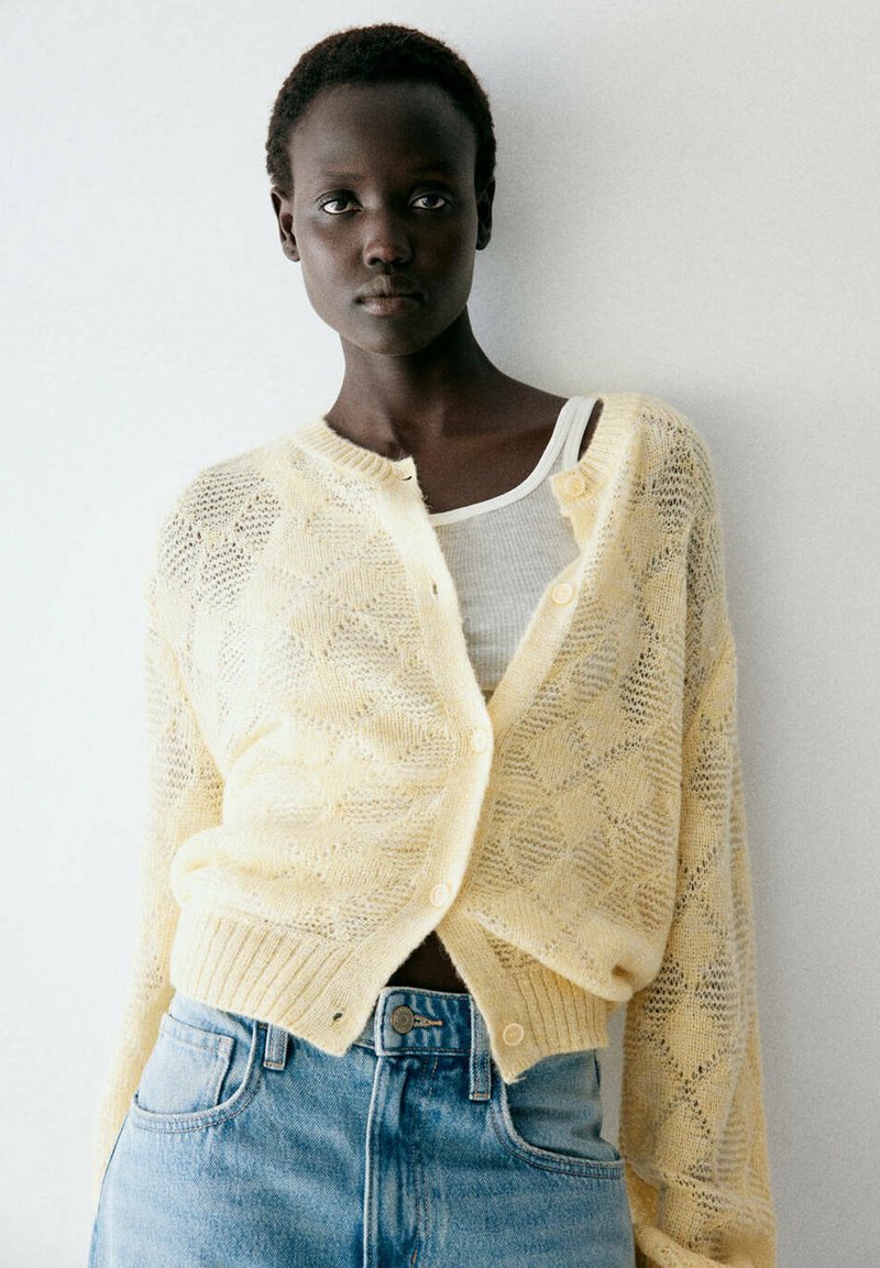 Femme aux cheveux courts portant un cardigan tricoté jaune pâle et un débardeur blanc, assortis à un jean en denim bleu clair sur un fond uni.