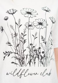 T-shirt blanche avec un design floral noir comprenant diverses fleurs sauvages et tiges, portant l'inscription "wildflower club" en lettres cursives en bas.