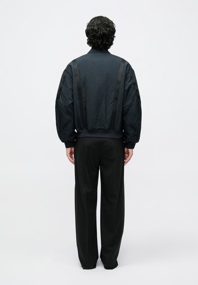 Čierna bomber bunda s rebrovaným golierom, nadrozmerným strihom, textúrovanou látkou a vertikálnymi panelmi. Kombinovaná s voľnými čiernymi nohavicami.