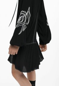 Schwarzes Kleid mit Puffärmeln, das mit weißer Blumenstickerei und einem plissierten Rock versehen ist. Weicher Stoff mit überlappenden Nahtdetails.