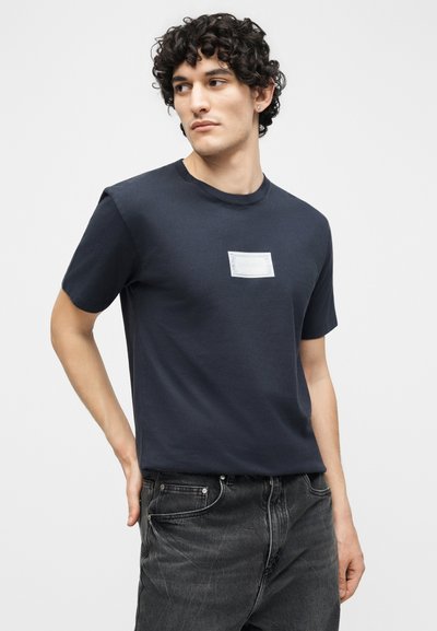 Νεαρός με σγουρά μαλλιά, φορώντας μπλε ναυτικό t-shirt με μικρό λευκό ορθογώνιο λογότυπο στο στήθος και μαύρα τζιν, κοιτάζοντας προς τα αριστερά.