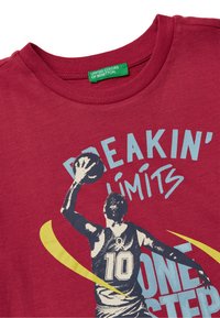 Červené bavlněné tričko má na sobě grafiku hráče, který hází basketbal a text "BREAKIN' LIMITS" v modrých a žlutých akcentoch. Velikost 10 je viditelná.