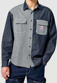Džínová košile na knoflíky se pruhem světle modré a bílé barvy na levé straně, tmavě modrou na pravé, se dvěma náprsními kapsami.