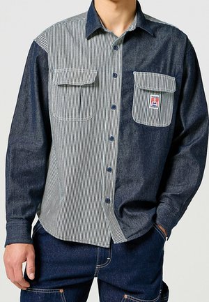 Camisa de mezclilla con botones que presenta un patrón a rayas en azul claro y blanco en el lado izquierdo, azul oscuro en el derecho, y con dos bolsillos en el pecho.