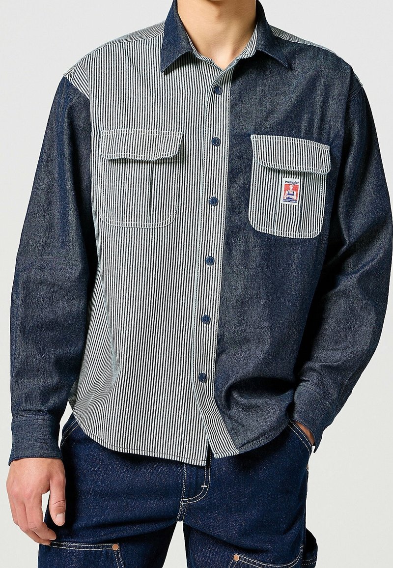 Džínová košile na knoflíky se pruhem světle modré a bílé barvy na levé straně, tmavě modrou na pravé, se dvěma náprsními kapsami.