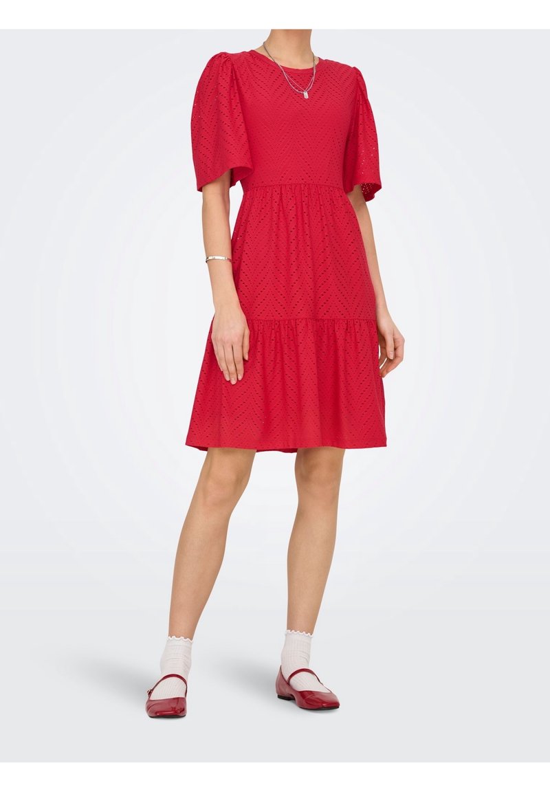 Femme portant une robe rouge longueur genou avec des manches bouffantes, des chaussettes blanches aux chevilles, des chaussures rouges Mary Jane et des bijoux en argent sur un fond uni.