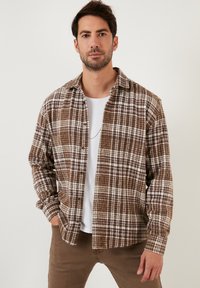 Camicia di flanella a quadri marrone e bianca, con colletto, bottone sul davanti, maniche lunghe, tessuto testurizzato, indossata sopra una t-shirt bianca.