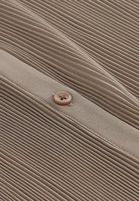 Plisseret stof i en dæmpet beige tone med en glat tekstur, prydet med en cirkulær brun knap og en subtil glans.