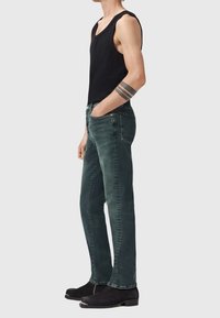 Muž v černém tílku, tmavě zelených džínách a černých botách, se pruhovaným tetováním na paži, stojí bokem s jednou rukou v kapse.