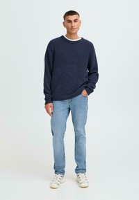 Tumši zils adīts džemperis ar apaļu kakla izgriezumu, kombinēts ar gaiši ziliem džinsiem un baltām sporta kurpēm ar melniem akcentiem.