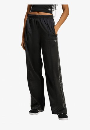 Persona che indossa pantaloni sportivi Adidas neri a gamba larga con strisce bianche laterali, top corto nero e sneakers nere, in piedi con una mano in tasca.