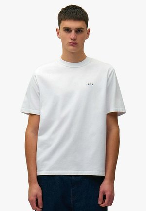 Giovane uomo che indossa una maglietta bianca a maniche corte con una piccola scritta nera "arte" sul petto a sinistra, abbinata a pantaloni blu scuro.