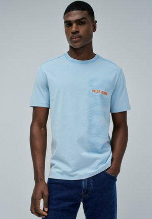Camiseta de algodón azul claro con cuello redondo, que presenta un gráfico texturizado en azul oscuro y un logo naranja de "Salsa Jeans" en el pecho.