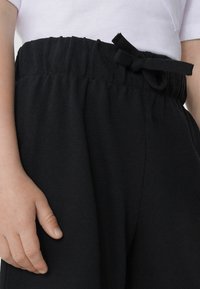 Pantalones de jogging noirs avec un tissu texturé, une taille élastique et un cordon ajustable. Une main repose sur la taille, mettant en valeur le design.