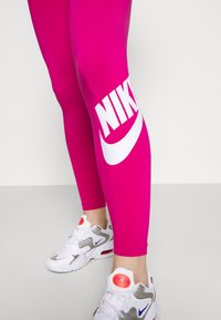 Leggings fucsia con grande logo bianco "NIKE" sulla parte bassa della gamba, abbinati a scarpe da ginnastica grigie, bianche e rosa neon.