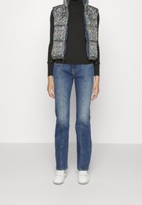 Kwiatowa kamizelka z wypełnieniem i falbaniastymi brzegami, czarny prążkowany golf, przyciemniane niebieskie jeansy o prostym kroju i białe sneakersy.