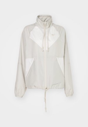 Giacca a vento grigio chiaro con accenti bianchi, colletto alto, vita regolabile con cordino, polsini elasticizzati e due tasche frontali. Logo Lacoste visibile.